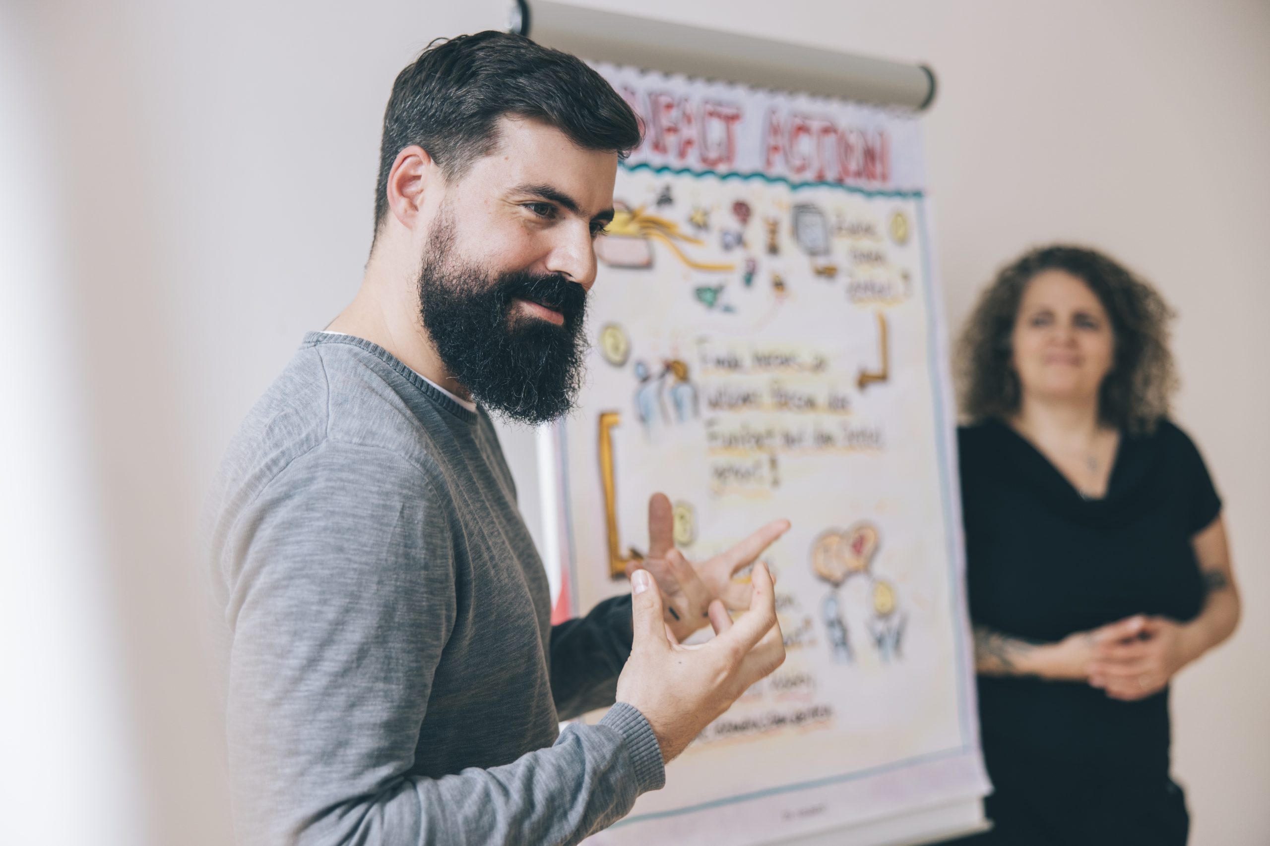 Auf dem Foto steht daniel Kauer, Trainer und Coach von COM ACROSS, neben einem Flipchart und gestikuliert begeistert und lächelnd, während einer Situation in einem Com-Across Workshop. Im Hintergrund befindet Fadja Ehlail und hört aufmerksam zu.