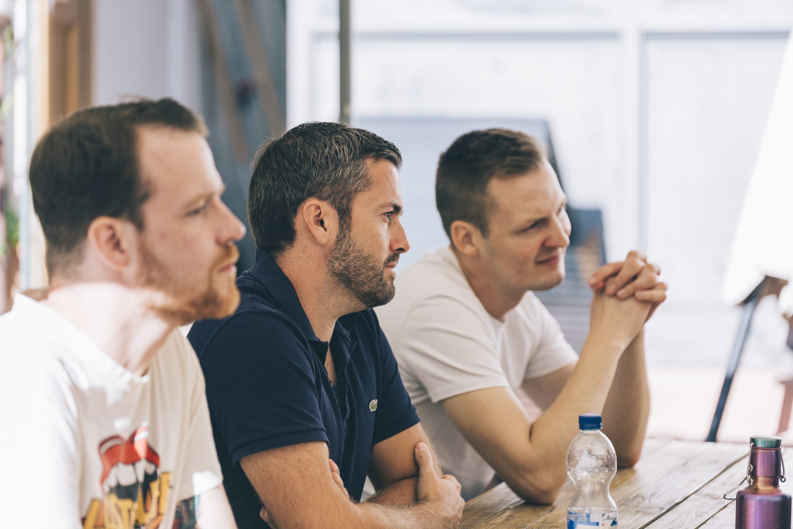 Auf dem Foto hören drei männliche Workshop-Teilnehmer aus dem Team von COM ACROSS aufmerksam und konzentriert zu.