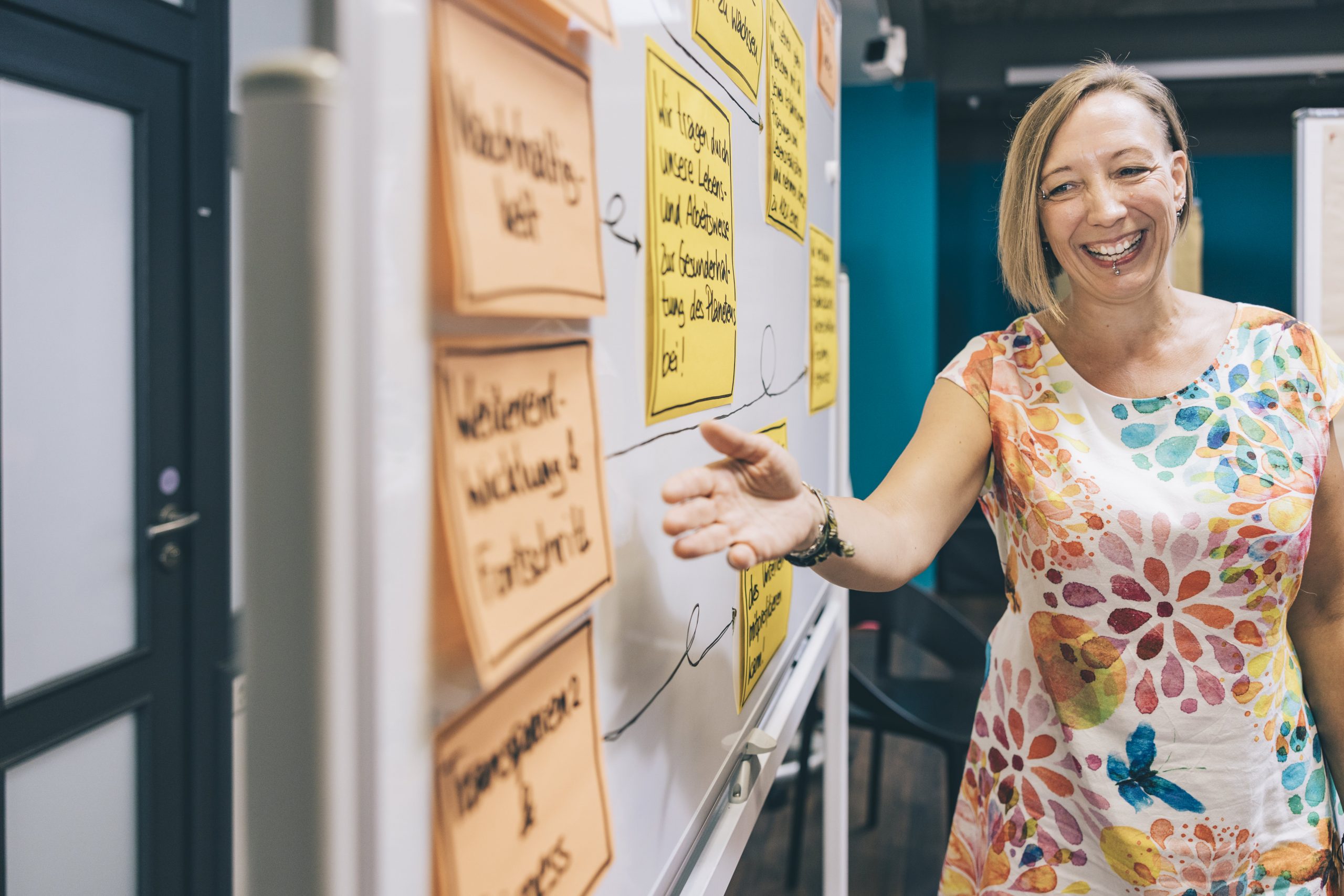 Auf dem Foto ist Heike Maerki aus dem Team von Com-Across in einer Workshopsituation abgebildet. Sie zeigt lachend auf einen Post-It auf einer Moderationswand.